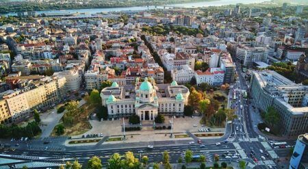 Aerial view of Belgrade, Serbia, showing the National Assembly building with green domes surrounded by dense cityscape, parks and a busy multi-lane road junction, with the Danube river visible in the background