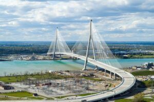 A wide aerial daytime view of the full cable-stayed bridge span with both towers and their cables, crossing the river between green riverbanks under a cloudy sky