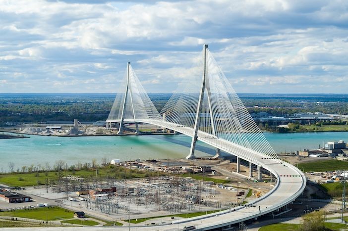 A wide aerial daytime view of the full cable-stayed bridge span with both towers and their cables, crossing the river between green riverbanks under a cloudy sky