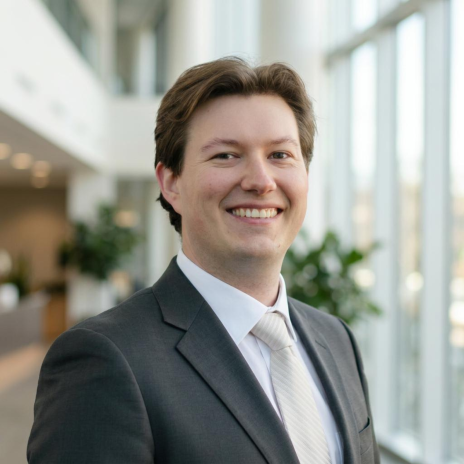 Headshot portrait of young man with center parted light brown hair smiling in a dark suit and tie