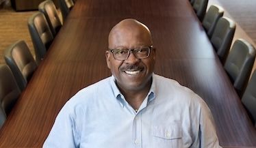 A bald man with glasses in an open necked blue shirt stands in front of a boardroom table with empty seats, smiling warmly