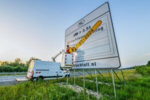 A worker in a high-visibility jacket, elevated on a cherry picker mounted on a white vehicle, removes a yellow date strip reading "01-07-2026" from a large Dutch roadside information sign. The sign displays a truck icon, the text "Vrachtwage..." and "trucktoll.nl" against a backdrop of motorway verge, trees and a pale early-morning sky.