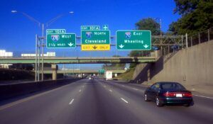 Overhead motorway signage on Interstate 70 directing traffic to I-670 West, I-71 North to Cleveland, and I-70 East to Wheeling, with a car passing beneath on a multi-lane highway