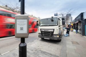 A "Southwark Loading Bays" signage pole with a QR code on a London street, with a red double-decker bus passing and a delivery lorry parked at the kerbside while a driver unloads crates onto the pavement
