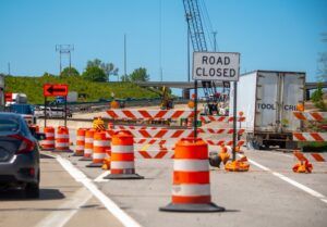 A "Road Closed" sign and orange-and-white traffic barrels lining a closed highway lane at a construction work zone, with a crane and trailer visible in the background