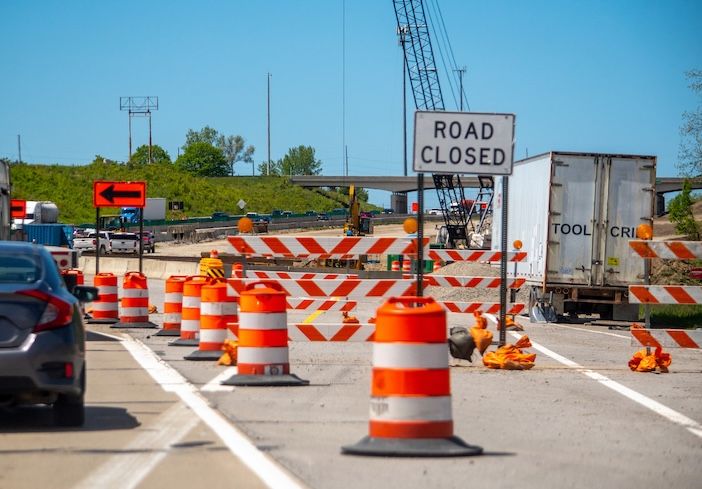A "Road Closed" sign and orange-and-white traffic barrels lining a closed highway lane at a construction work zone, with a crane and trailer visible in the background
