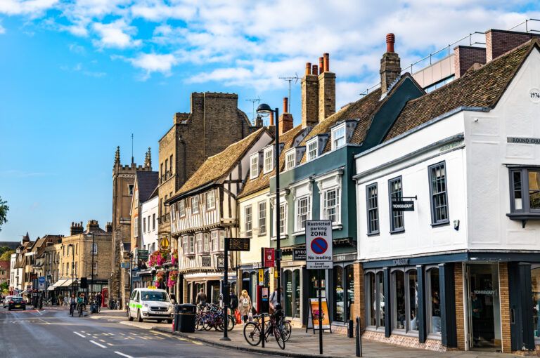 A sunny street scene in central Cambridge showing historic buildings, shops, cyclists and pedestrians along Bridge Street.