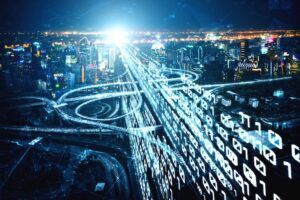 Aerial night-time view of a motorway interchange overlaid with glowing streams of binary code, representing digital traffic data flowing across a road network