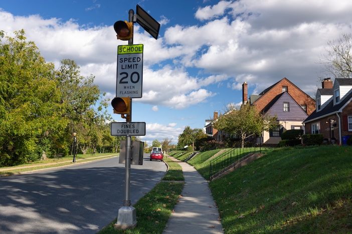 Quiet suburban street in Frederick, MD, with two-story houses, green lawns, and a school zone warning sign flashing at 20 miles per hour, emphasizing the importance of road safety