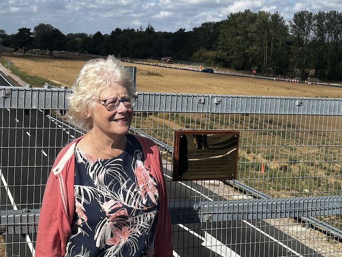 Carol Charles, daughter of road safety campaigner Valerie Knights MBE, stands beside a commemorative plaque on the newly built bridge over the A47, with the new dual carriageway and roadworks visible in the fields behind her