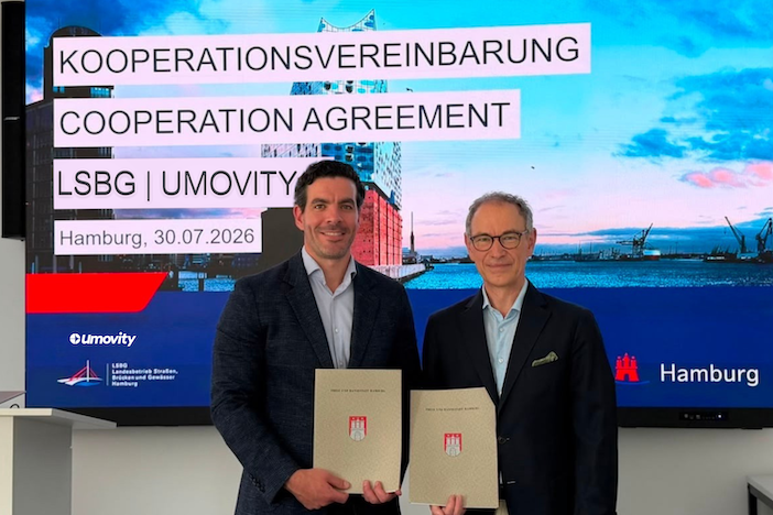 Christoph Heel and Nick Ghia stand side by side holding document folders in front of a large screen displaying the text "Kooperationsvereinbarung / Cooperation Agreement, LSBG | Umovity, Hamburg, 30.07.2026" with the LSBG and Umovity logos and a Hamburg skyline image behind them