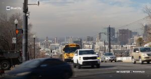 A pole-mounted lidar sensor beside a pedestrian crossing signal on a Salt Lake City street, with traffic including a school bus and the city skyline in the background.
