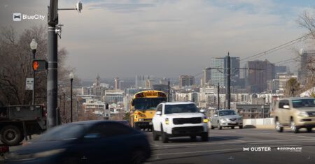 Econolite wins UDOT deal to extend Ouster BlueCity to 160 intersections A pole-mounted lidar sensor beside a pedestrian crossing signal on a Salt Lake City street, with traffic including a school bus and the city skyline in the background.