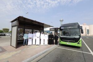 Officials from the Integrated Transport Centre and the Authority of Social Contribution – Ma'an, including women in traditional black abayas and men in white kanduras, pose beside one of the new bus shelters and a green Abu Dhabi Mobility electric bus