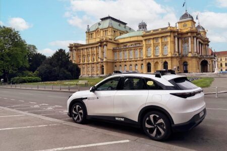 Pony.ai and Verne launch driverless robotaxi tests in Zagreb, Croatia A white SUV with a sensor pod on its roof and "verne" branding on its door is parked in a plaza in front of an ornate yellow domed building, with flagpoles and trees nearby on a sunny day