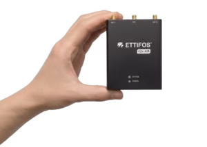A hand holds a small black rectangular electronic device labelled Ettifos V2X-AIR, showing three antenna connectors marked ANT1, GPS and ANT2 along the top edge and two indicator lights labelled STATUS and POWER on the front panel.