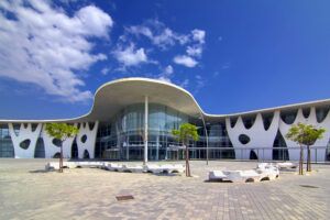 A modern white exhibition building with a wavy overhanging roof and a glass entrance, fronted by a paved plaza with young trees and curved stone benches under a blue sky