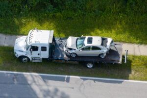 erial view of a flatbed tow truck carrying a crash-damaged silver sedan with a caved-in roof and shattered windows along a roadside verge