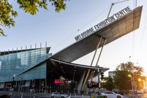 large building with an angular, slanted metal roof reading 'Melbourne Exhibition Centre' in bold letters, seen from street level at sunset, with tram warning signs, traffic lights and pedestrian crossings in the foreground and tree branches overhead."