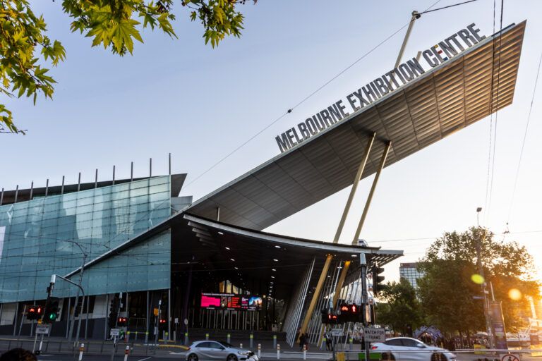 large building with an angular, slanted metal roof reading 'Melbourne Exhibition Centre' in bold letters, seen from street level at sunset, with tram warning signs, traffic lights and pedestrian crossings in the foreground and tree branches overhead."