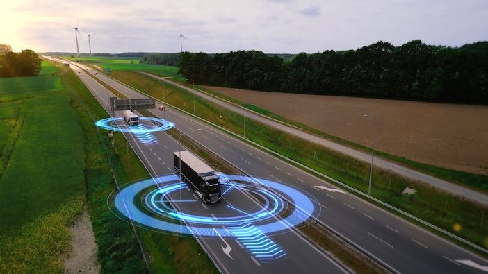 Aerial view of two trucks on a highway at dusk, each surrounded by glowing blue circular sensor graphics, with wind turbines and green fields in the background