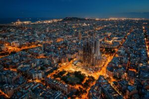 Aerial night-time view of Barcelona, with the illuminated Sagrada Família basilica standing above the surrounding city grid and the coastline visible in the distance