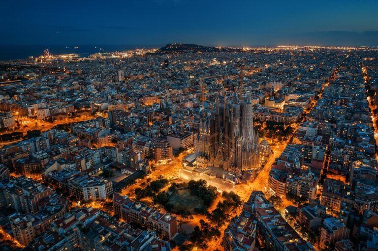 Aerial night-time view of Barcelona, with the illuminated Sagrada Família basilica standing above the surrounding city grid and the coastline visible in the distance