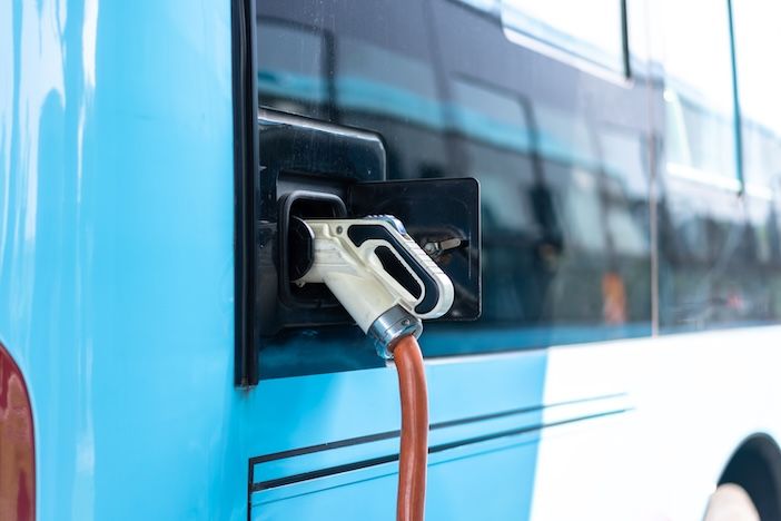 Close-up of a charging connector plugged into a socket on the side of a light blue bus, with an orange charging cable running down to the ground