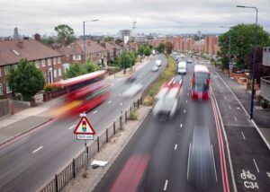 Elevated view of a dual carriageway lined with red-brick houses, with motion-blurred red double-decker buses and cars, a traffic signals warning sign and a painted cycle lane in the foreground