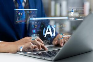 A person typing on a laptop with holographic AI dashboard graphics overlaid, including charts, a chatbot icon and an "Ai" symbol, representing artificial intelligence tools