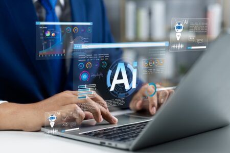 A person typing on a laptop with holographic AI dashboard graphics overlaid, including charts, a chatbot icon and an "Ai" symbol, representing artificial intelligence tools