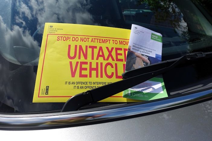 bright yellow notice reading "STOP! DO NOT ATTEMPT TO MOVE, UNTAXED VEHICLE" is tucked under a car's windscreen wiper, next to a leaflet about wheel-clamping and removing vehicles, with trees and sky reflected in the windscreen