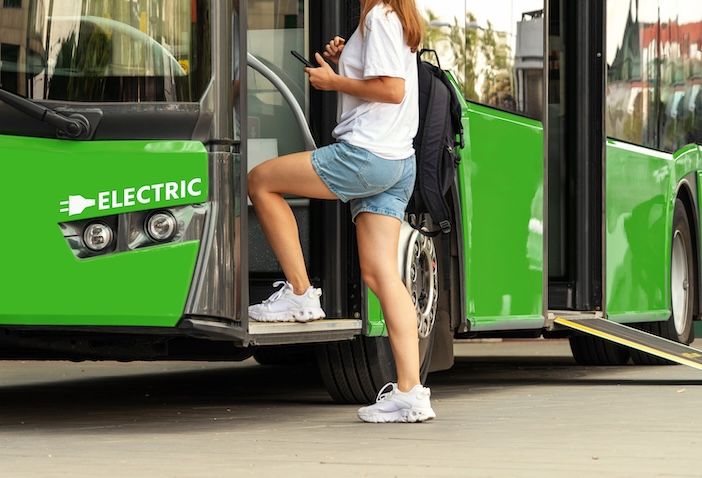 A woman carrying a backpack and looking at her phone boards a green bus marked "ELECTRIC" via a low-floor ramp