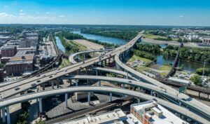 Aerial view of a multi-level elevated highway interchange with curving ramps and traffic, beside a river lined with trees and city buildings under a blue sky
