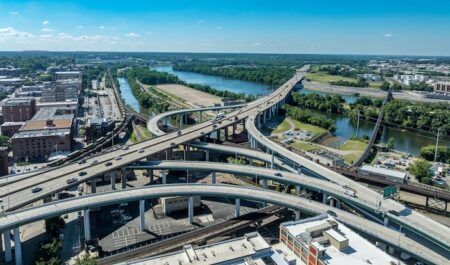 Aerial view of a multi-level elevated highway interchange with curving ramps and traffic, beside a river lined with trees and city buildings under a blue sky