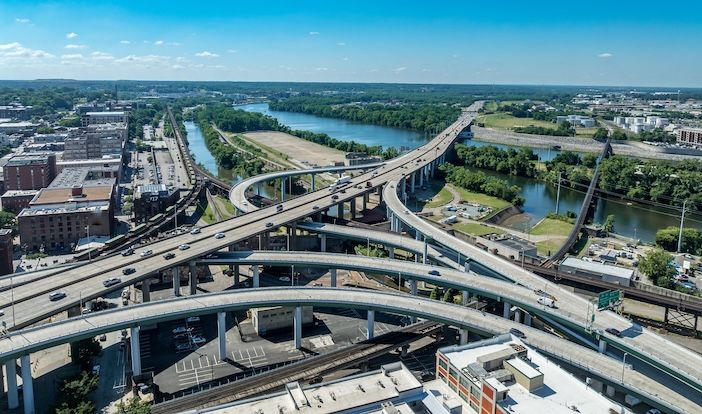 Q-Free launches cybersecure Dynamic Metering for freeway ramps Aerial view of a multi-level elevated highway interchange with curving ramps and traffic, beside a river lined with trees and city buildings under a blue sky