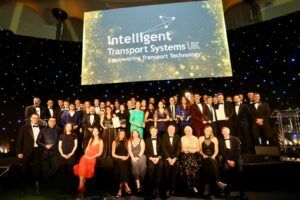 A large group of people in formal evening wear, many holding glass trophies and certificates, pose on a stage beneath a screen showing the Intelligent Transport Systems UK logo