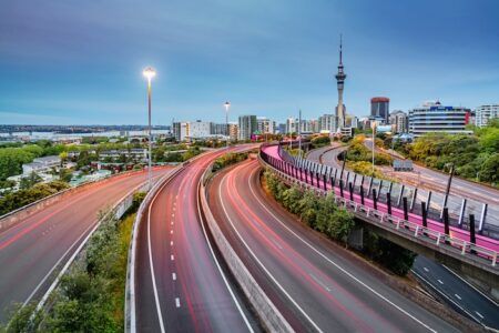 Kapsch TrafficCom upgrades New Zealand traffic system Auckland Sky Tower and motion blurred highway traffic lights during twilight. Illuminated Light Path Bicycle Lane beside the Highway. Auckland City, New Zealand.