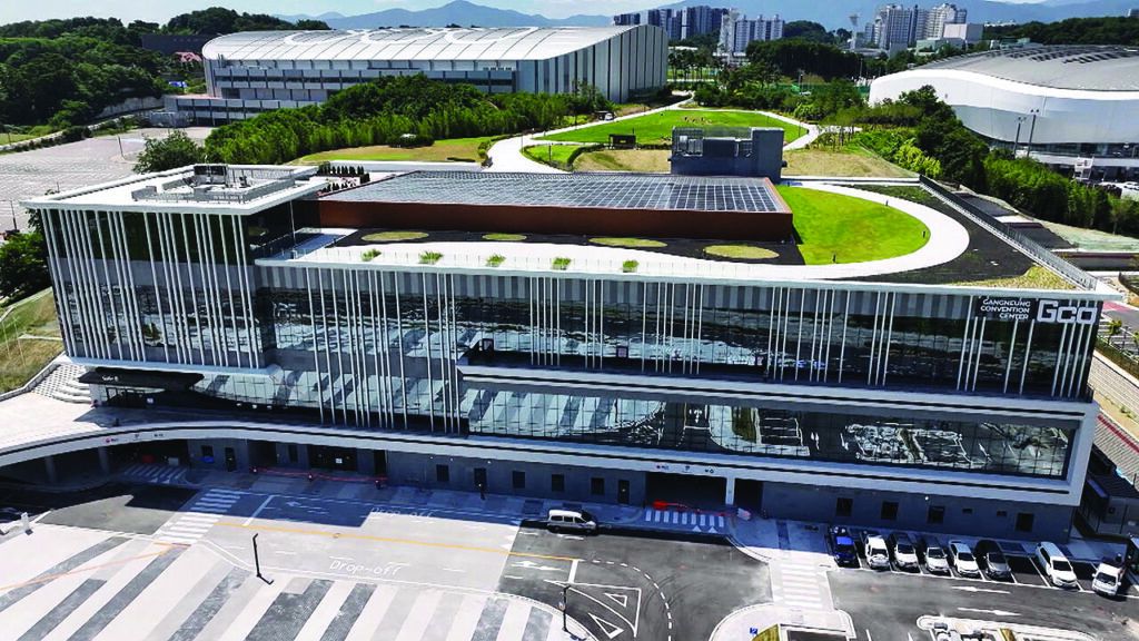 Aerial view of a modern glass-fronted convention centre with a green roof garden and solar panels, a drop-off area and car park in front, and other buildings visible behind