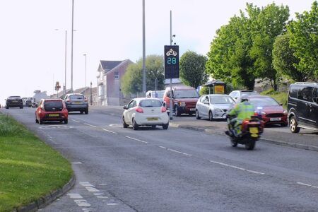 A vehicle activated sign at the roadside in Swansea, displaying a speed reading and an "ARAF SLOW" warning to an approaching motorcyclist