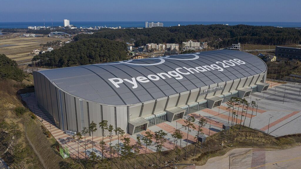 Aerial view of a large arena with a curved roof reading "PyeongChang 2018" beside the Olympic rings, surrounded by trees and a car park, with a coastline in the distance