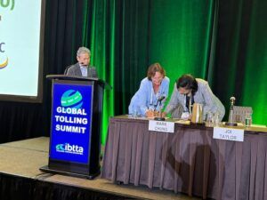 Two women lean over a draped table on a stage to sign documents while a man stands at a lectern displaying a Global Tolling Summit sign against a green curtain