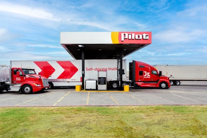 A red truck marked "self-driving truck" and branded with the Kodiak logo, parked under the fuel canopy of a Pilot truck stop, with a second truck and trailer parked alongside