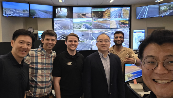 A group of six men pose for a selfie-style photo in a traffic management control room, with a wall of monitors displaying live highway and intersection camera feeds behind them.