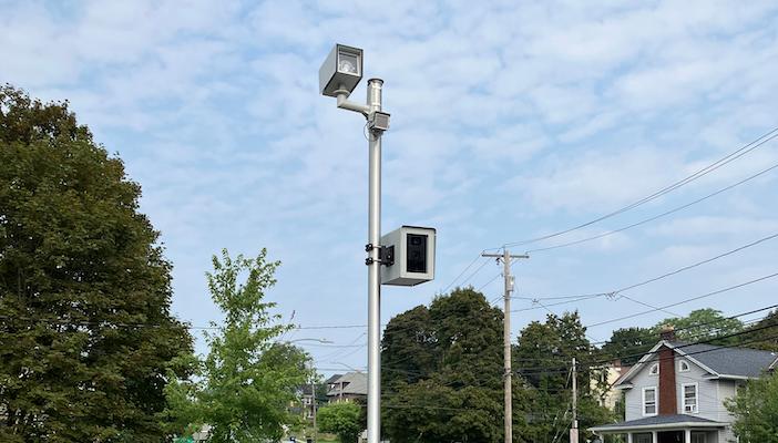 Jenoptik wins deal for 140+ intersection safety cameras in Canada A tall metal pole beside a suburban road with two box-shaped cameras mounted on it and a grey cabinet near the base, with houses, trees and a passing car in the background