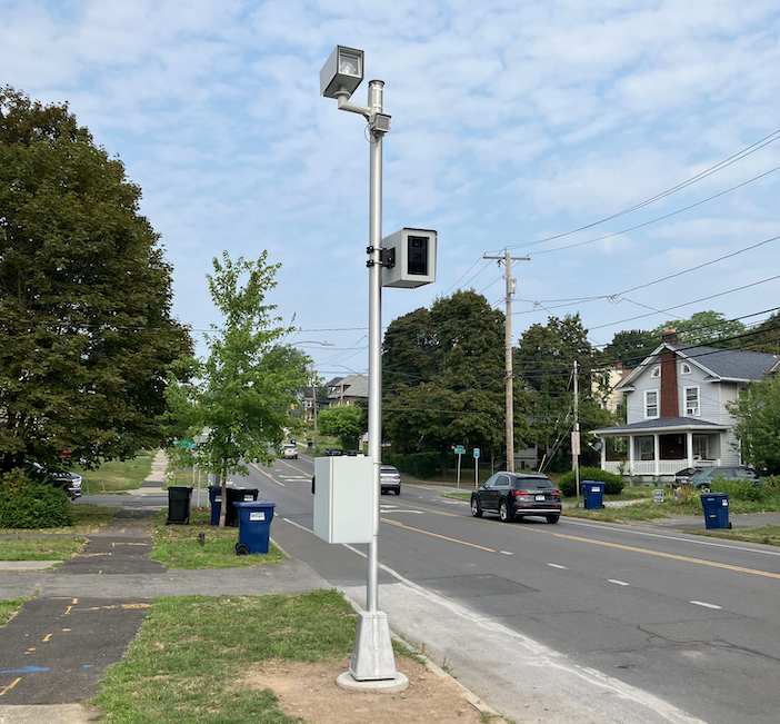 A tall metal pole beside a suburban road with two box-shaped cameras mounted on it and a grey cabinet near the base, with houses, trees and a passing car in the background