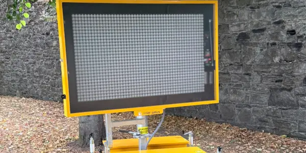 A yellow-framed LED variable message sign display mounted on a compact base unit, standing against a stone wall surrounded by fallen autumn leaves