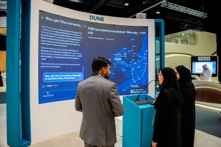 A man in a grey suit and two women in black abayas stand in front of a large illuminated screen showing a chat interface and a blue metro network map, with a lectern and microphone beside them
