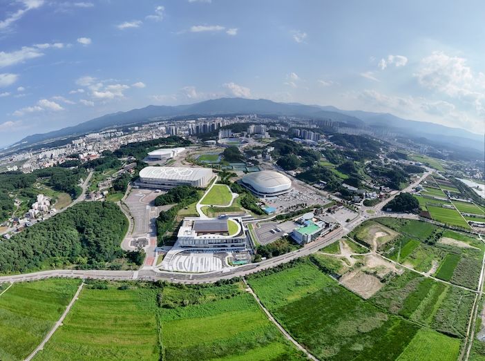 Wide-angle aerial view of the conference centre complex and surrounding arenas, set among green fields and hills, with a town and mountains visible in the distance