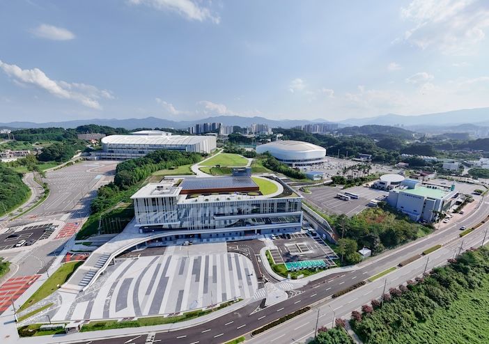 Aerial view of a modern conference centre with a sloped green roof and solar panels in the foreground, surrounded by car parks and two large white domed arena buildings, with hills and a town beyond